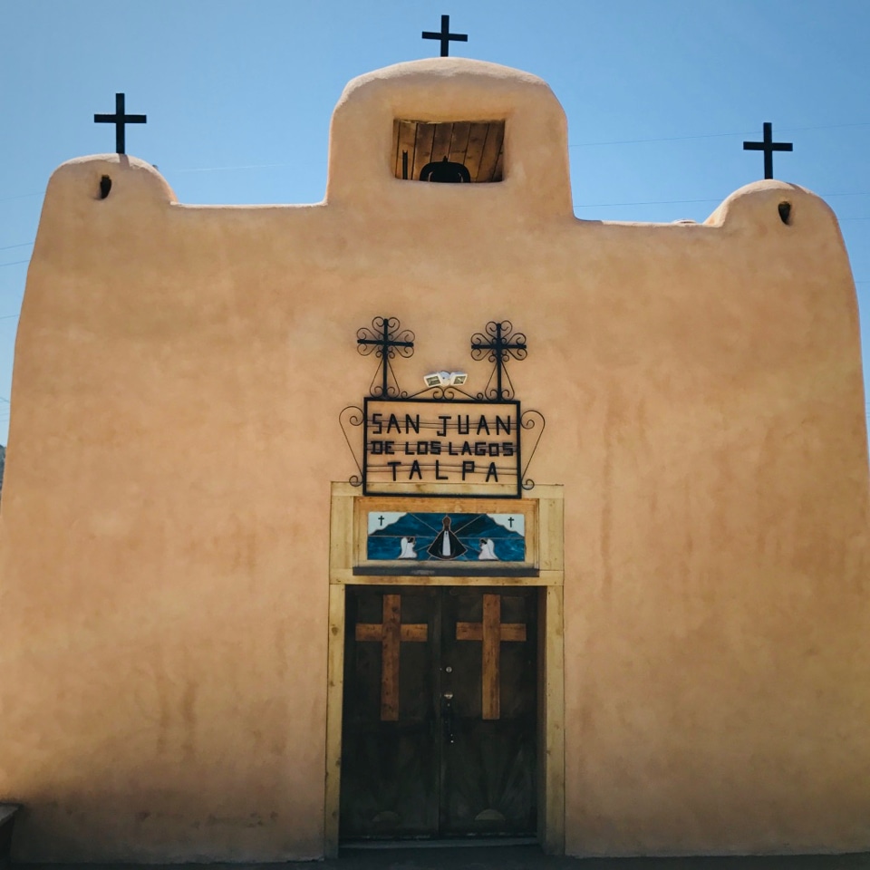 A small adobe church with a bell tower and three crosses on top, and a sign reading San Juan de los Lagos Talpa