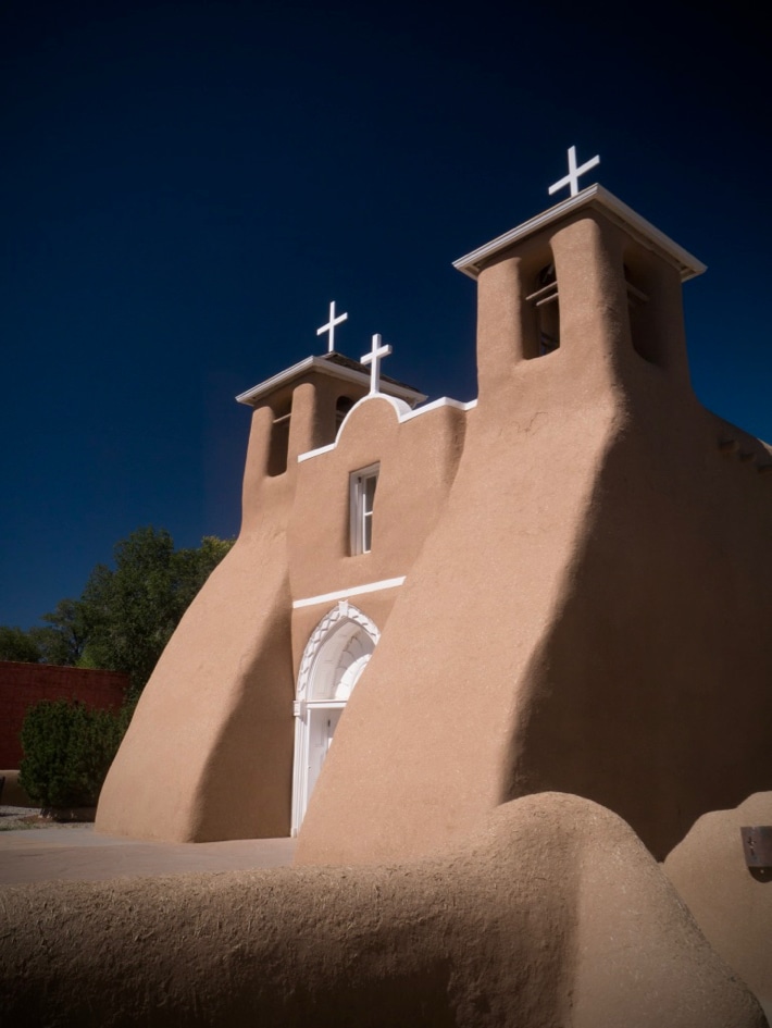 An adobe church, under a blue sky, with white wooden door and crosses, and two bell towers