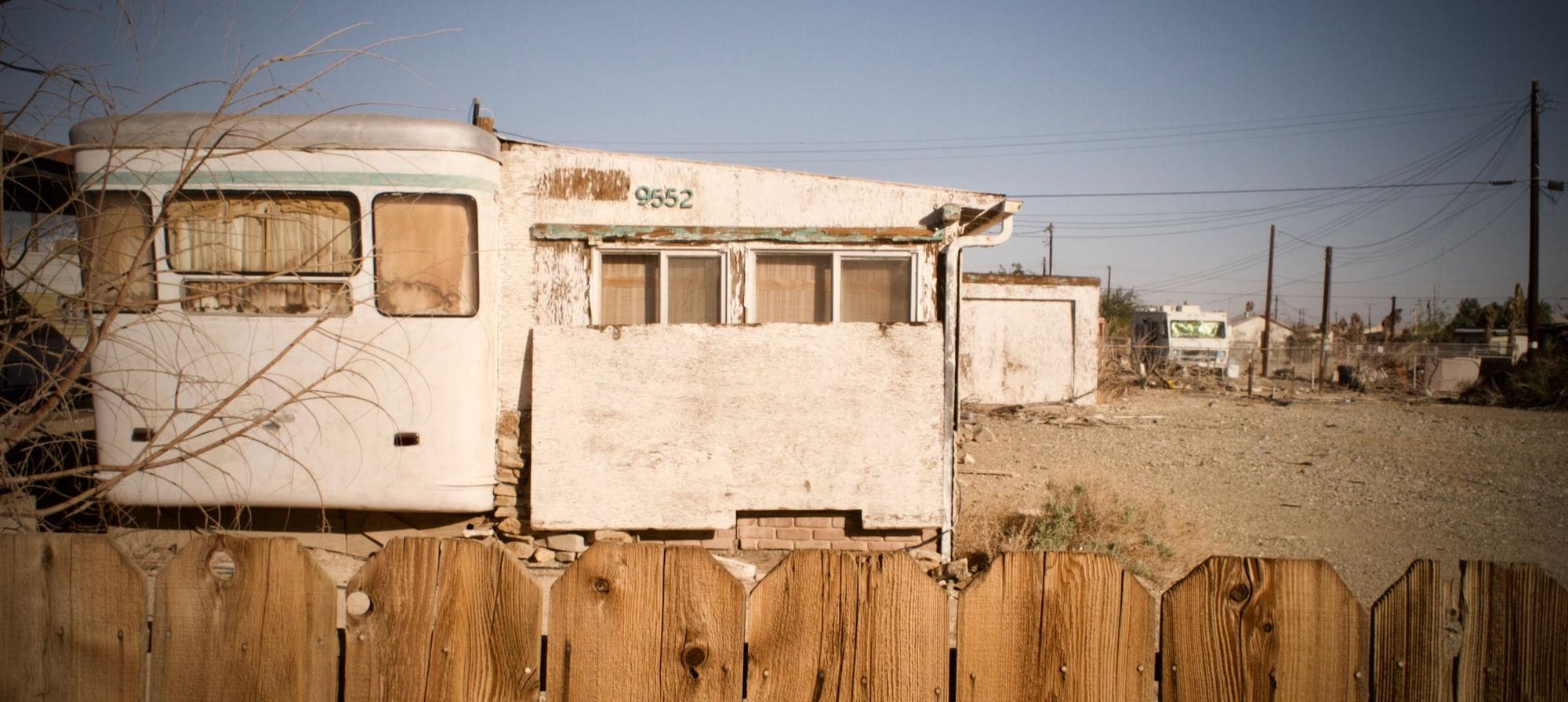 An abandoned old mobile home in the desert, with a wooden fence and dried-out tree in front of it