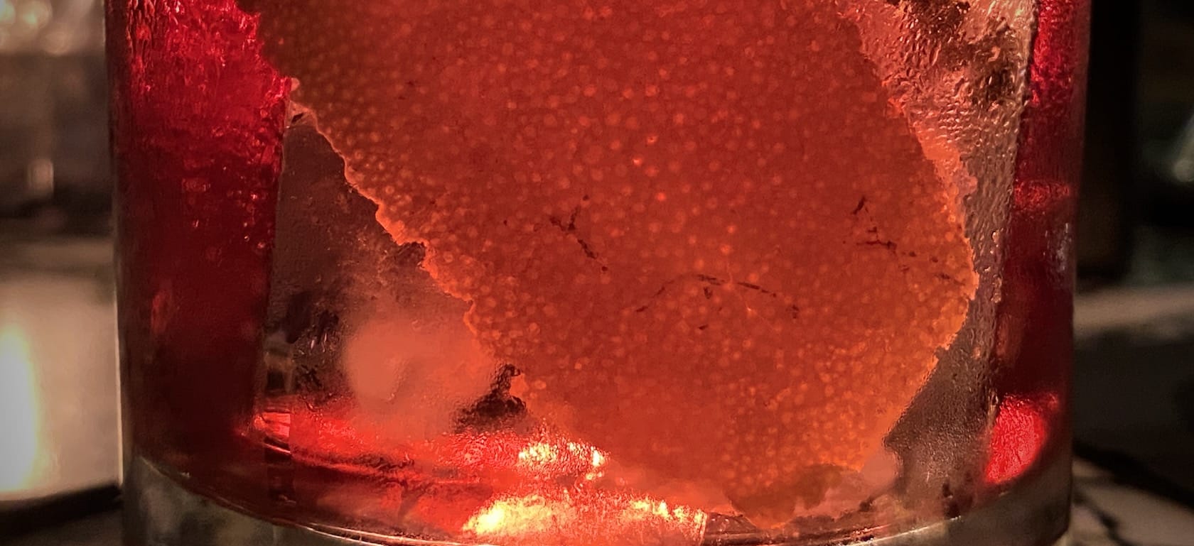 A closeup on a sweating glass, icecubes, and an orange liqueur in it -- a Negroni