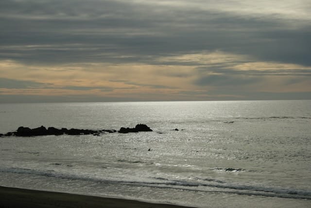 The sea in afternoon, a shiny gray surface under gray clouds, some gold from sunset creeping in.