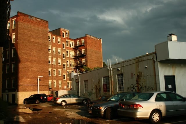 A brick apartment building and parking lot glowing golden in the afternoon sun, with a cloudy gray sky above. Many cars parked in the wet, potholed lot. 